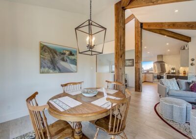 Open-plan living space with a wooden dining table, coastal artwork, and exposed beams, leading into a kitchen area.