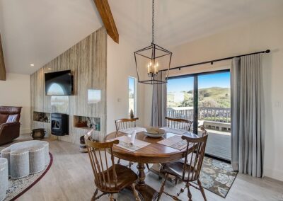 A contemporary dining room with a wooden table set, a fireplace, exposed wooden beams, and a view of the hills through the sliding glass door.