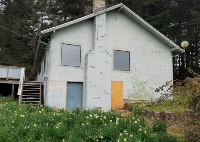 Dilapidated two-story house with peeling paint and boarded-up windows surrounded by overgrown greenery.